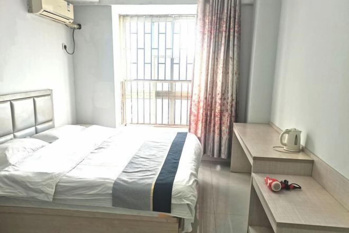 Ein Schlafzimmer mit einem Bett, einem Nachttisch mit Wasserkocher und einem roten Gegenstand, einem Fenster mit Gitter und einem Vorhang.