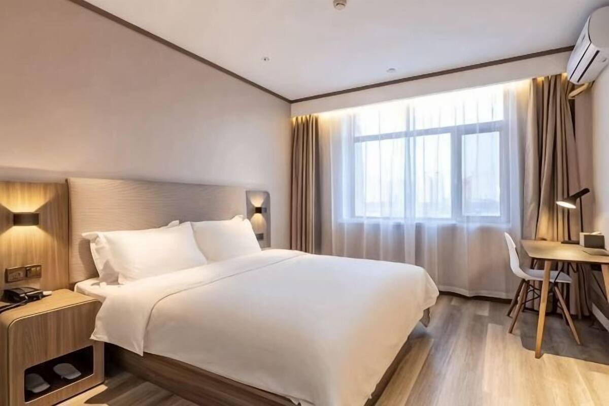 Habitación de hotel con cama, mesita de noche, escritorio, silla y ventana grande con cortinas.