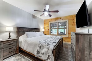 Une chambre à coucher avec une tête de lit en bois, un lit recouvert d’une couverture à motifs blancs et gris, une table de chevet avec une lampe, une commode et un ventilateur de plafond.