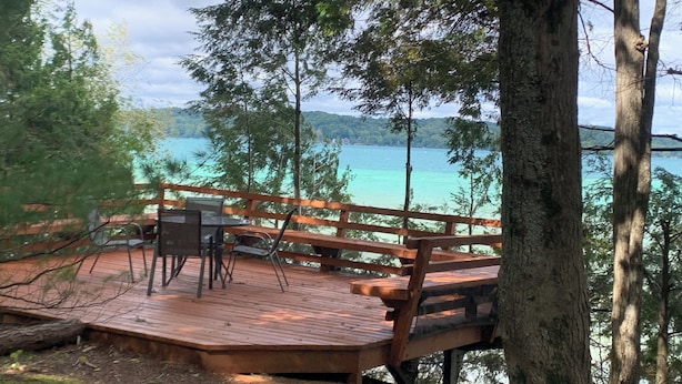 A wooden deck with a table and chairs overlooking a turquoise lake. - Torch lake waterfront:100 ft of sandy beach, 3-bedroom, 3-bath cottage (Alden)