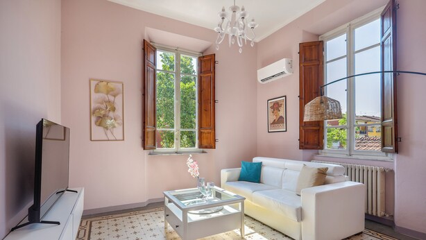 Uma sala de estar com um sofá branco, uma mesa de centro de vidro, uma TV sobre um suporte e uma janela grande com persianas de madeira.