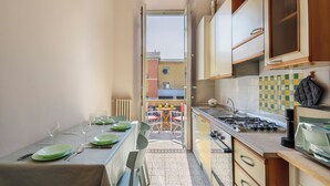 Cozinha compacta com área de refeições, vista através de uma porta aberta para uma área externa com assentos e painel de azulejos.