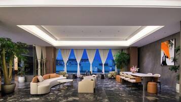 A spacious lobby with a modern design, featuring a seating area with sofas and a long table, a large window with curtains, and potted plants.