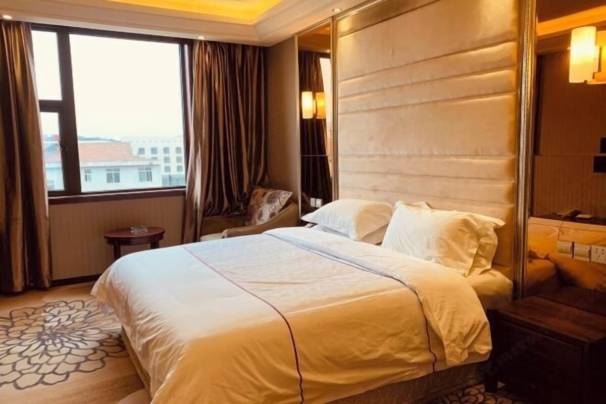 A hotel room with a large bed, a small side table, a chair, a mirror, and a view of buildings through the window.