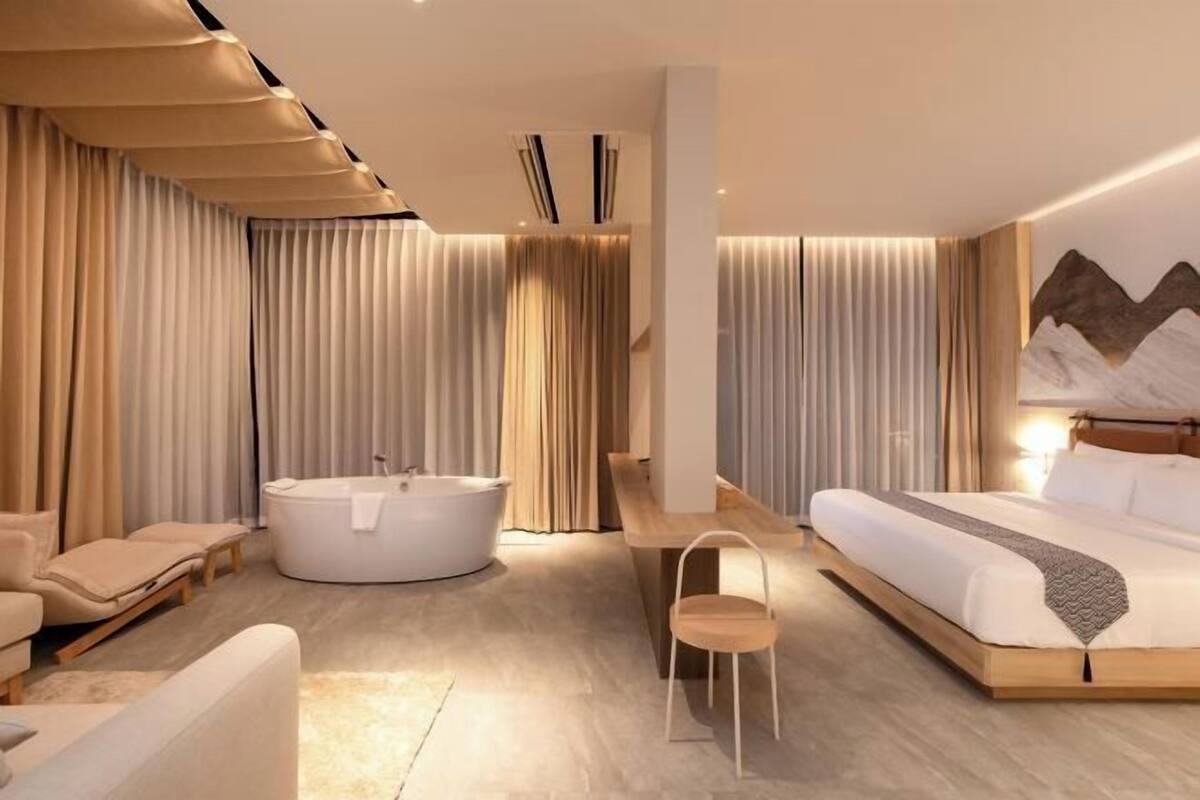 A modern hotel room with a large bed, a bathtub, a seating area, and a wall-mounted artwork.