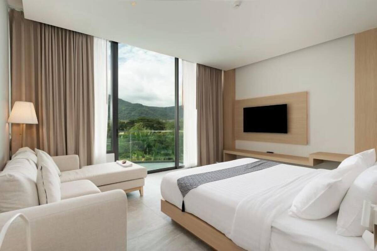 A modern hotel room with a large bed, a sofa, a TV, and a view of greenery through large windows.
