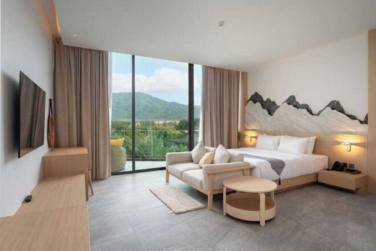 A modern hotel room with a large bed, a sofa, a small table, a TV, and a view of greenery and mountains.