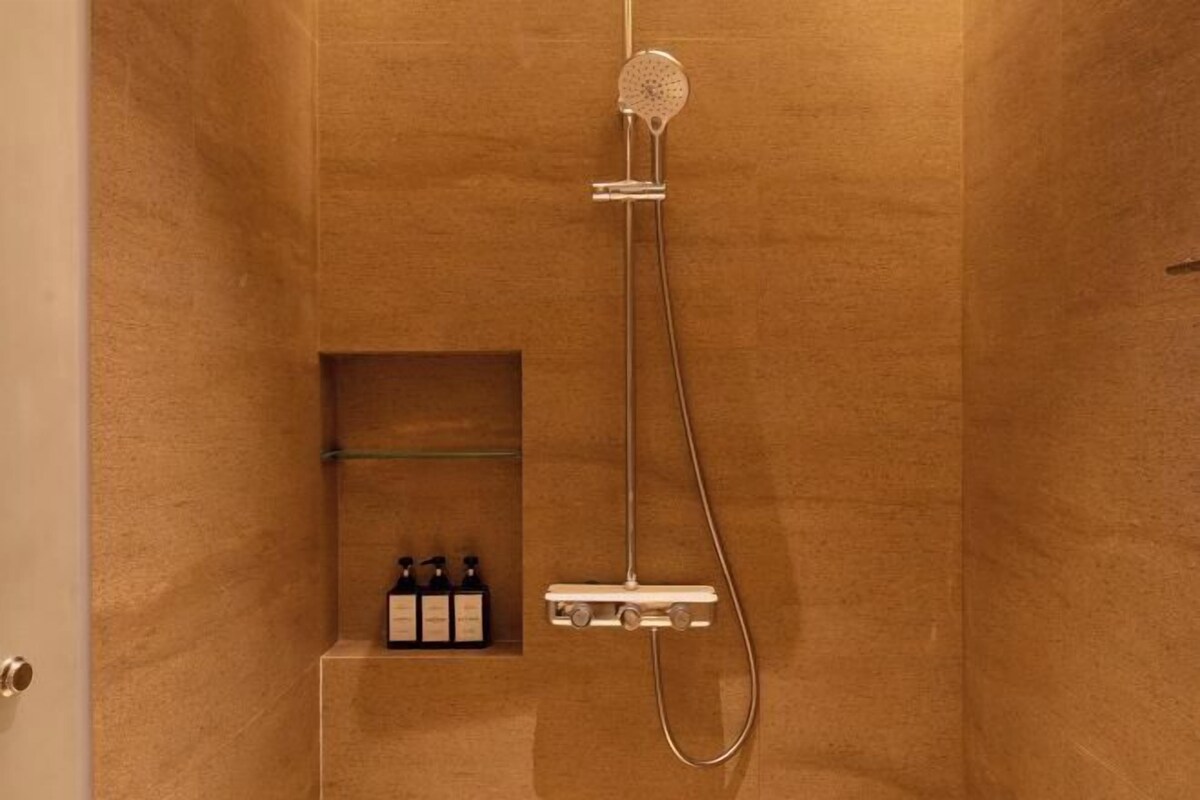 A shower with a handheld showerhead, a wall-mounted control panel, and a shelf with toiletries.