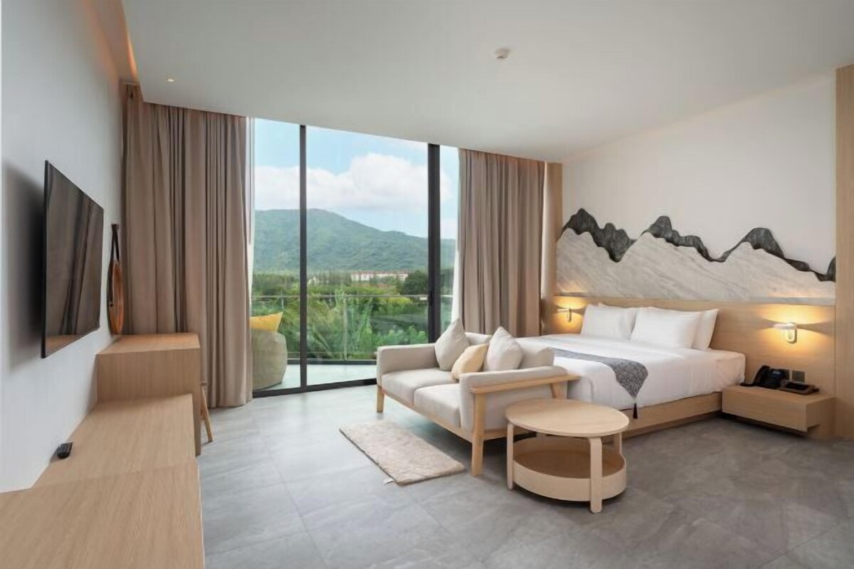 A modern hotel room with a large bed, a sofa, a small table, a TV, and a view of greenery and mountains.