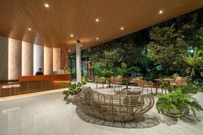 Un espace d'accueil doté d'un comptoir de réception en bois, d'un coin salon circulaire en osier et d'un jardin intérieur luxuriant agrémenté de diverses plantes et de différents types de sièges.