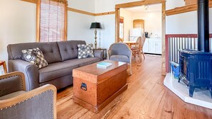 A cozy living room with a sofa, armchair, and a wooden coffee table. A blue wood-burning stove is visible in the background. - Stunning Cottage across for public Bayshore beach (Grand Marais)