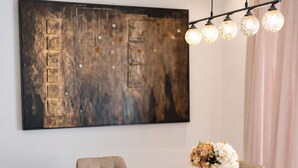 Un comedor con una mesa de mármol, sillas acolchadas, un cuadro abstracto grande y una lámpara colgante.