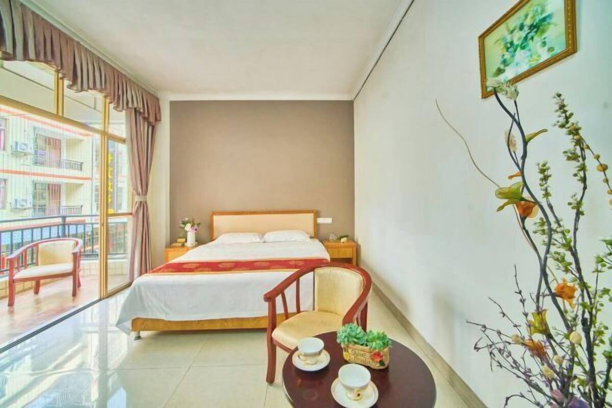 A hotel room with a bed, a chair, a small table with a cup and saucer, and a window with curtains.