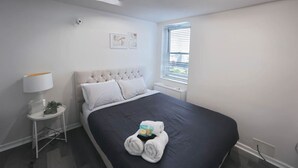 A modern bedroom with a large bed, a bedside table with a lamp, and a window with blinds.
