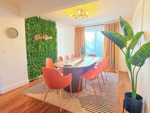 Une salle à manger avec une table ronde, des chaises orange, un mur vert orné de texte, une plante en pot et une fenêtre avec des rideaux.
