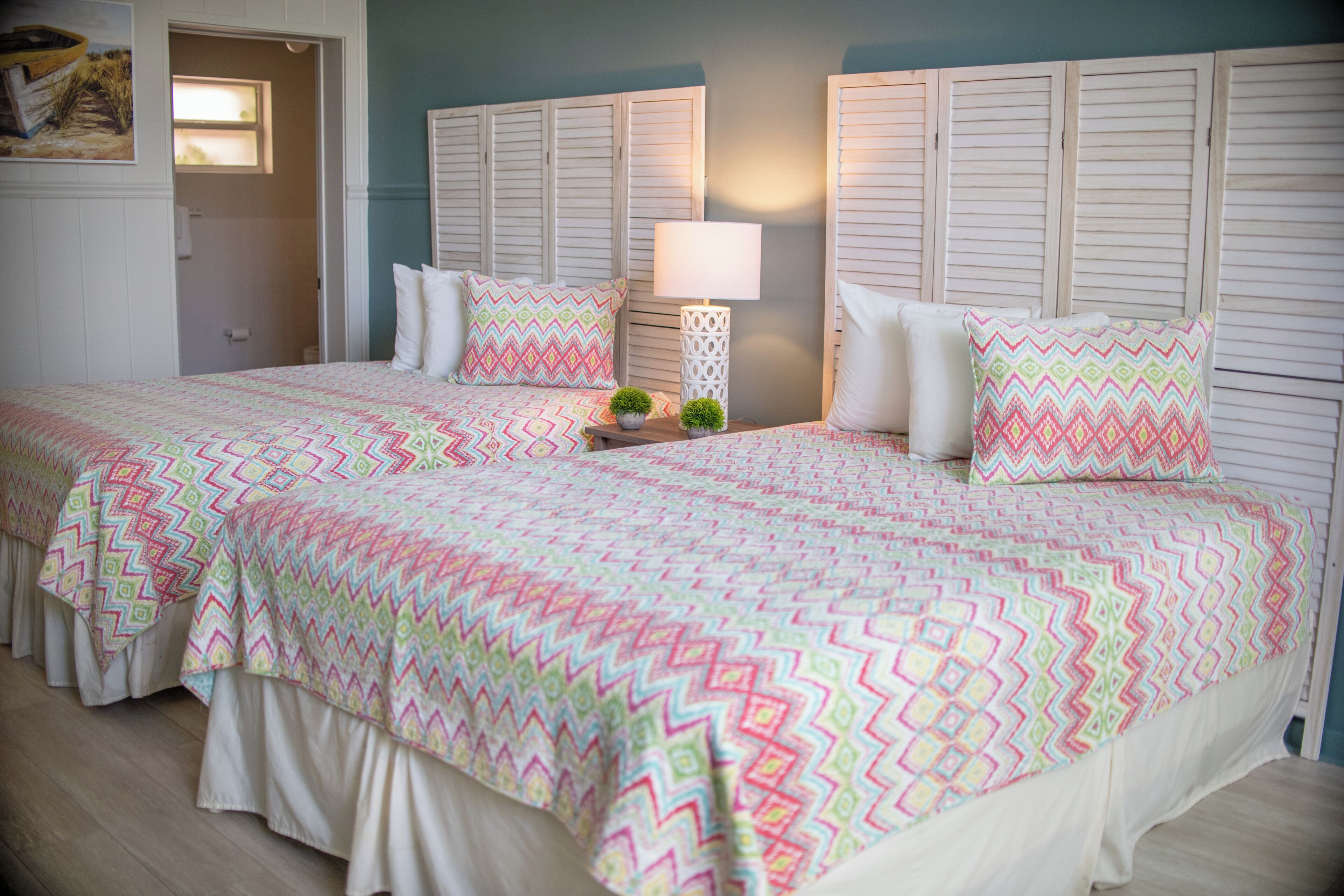 A double bed with a colorful zigzag patterned bedspread, two bedside tables with lamps, and a door leading to a bathroom.