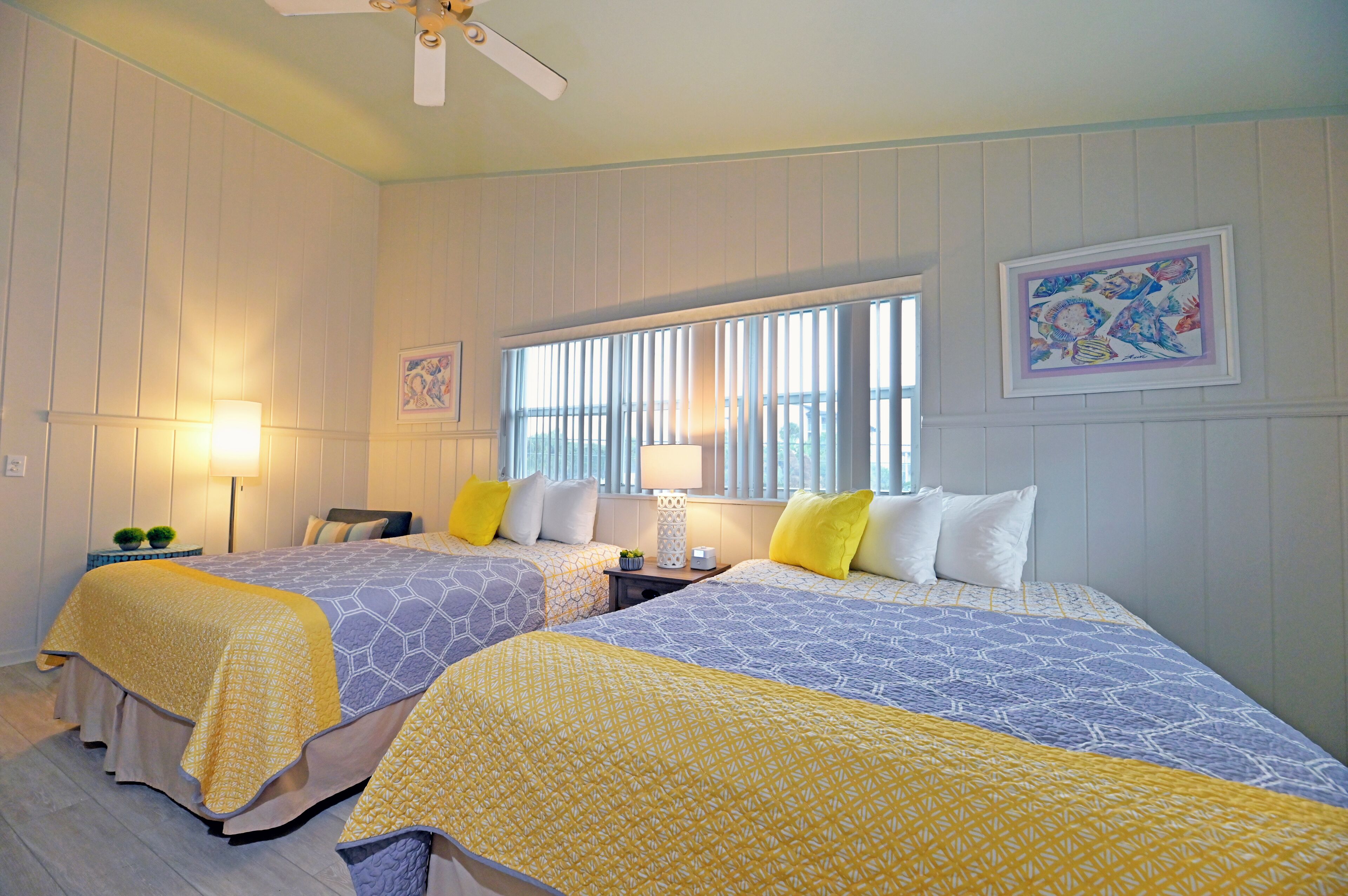 A hotel room with two beds, each with a patterned bedspread, a window with blinds, a ceiling fan, and a framed picture on the wall.