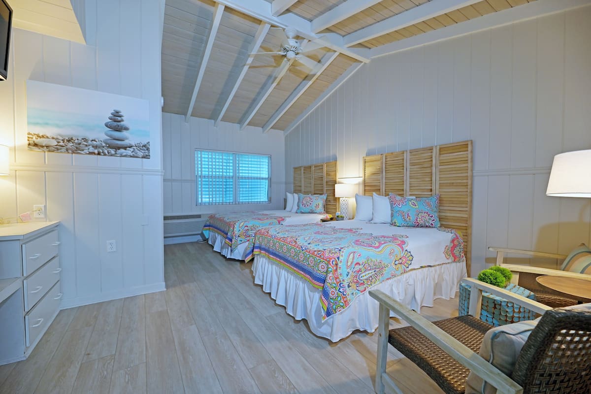 A bedroom with two beds, a ceiling fan, a window with blinds, and a bedside table with a lamp.