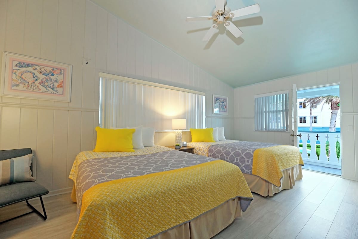A hotel room with two beds, a ceiling fan, and a balcony with a view of palm trees.