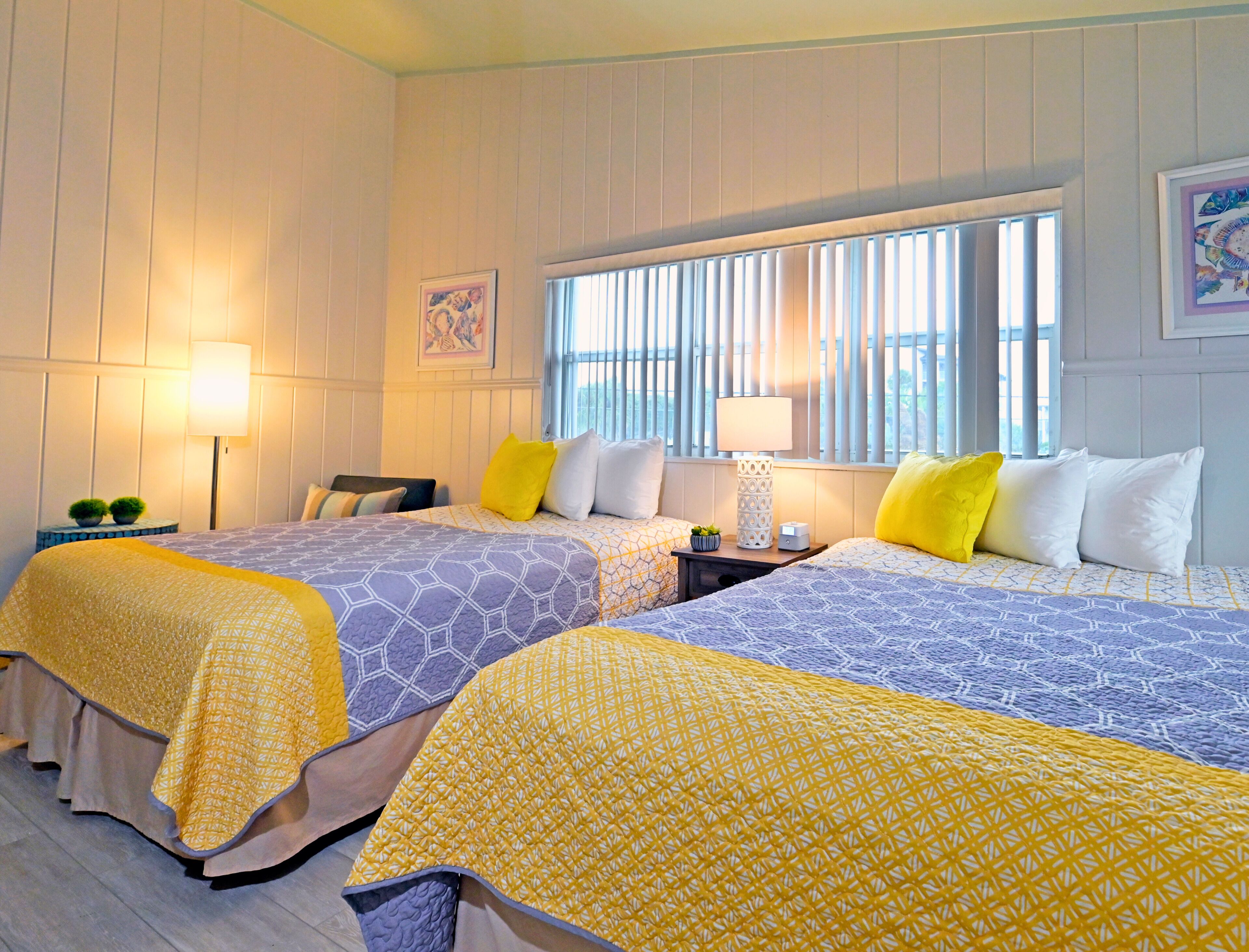 A hotel room with two beds, each with a patterned bedspread, a nightstand with a lamp, and a window with blinds.