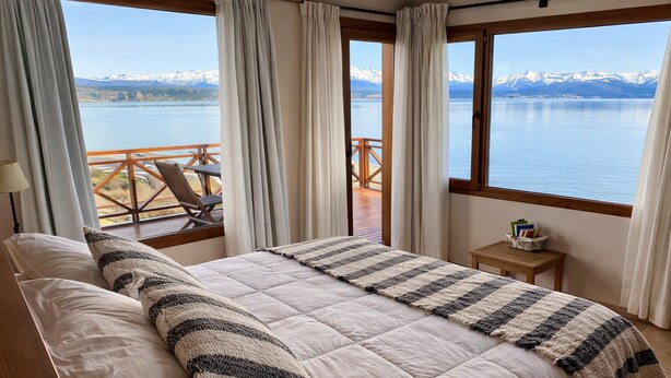 A bedroom with a large bed, a view of a lake and mountains, and a balcony with a wooden railing. - Crystal Suite Loft 2nd Floor Lake Front Bariloche (Dina Huapi)