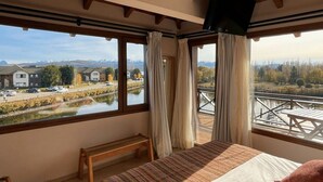 A bedroom with a large bed, wooden beams, and a view of a lake and buildings. - Amancay Lagoon Suite Loft 2nd Floor Lagoon Front (Dina Huapi)