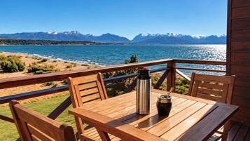 Une terrasse en bois avec une table et des chaises, donnant sur un lac et offrant une vue sur des montagnes en arrière-plan.