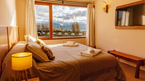 A hotel room with a bed, a bedside lamp, a TV, a window with a view of mountains and a lake, and a wooden bench. - Amancay 2nd Floor Lagoon & Bariloche View (Dina Huapi)