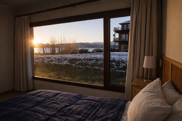 A bedroom with a large window offering a view of a snowy landscape and a building across the water.