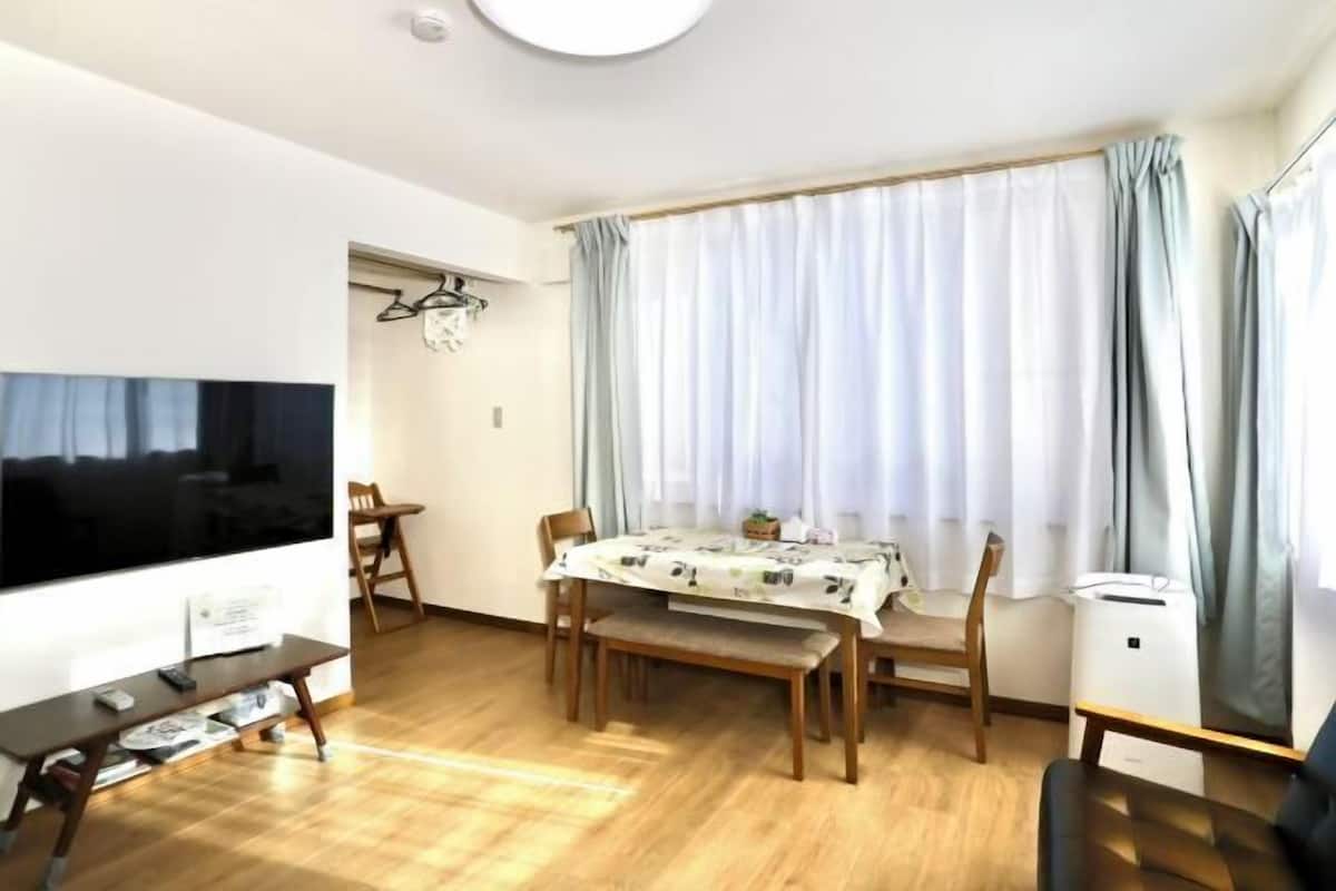 Ein Wohnzimmer mit einem Holzesstisch, Stühlen, einer Bank, einem Fernseher und einem Fenster mit Vorhängen.
