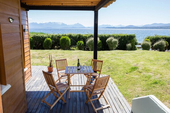A wooden deck with a table and chairs, overlooking a body of water and mountains.