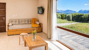A room with a sofa, a wooden table with a vase of flowers, a television on the wall, and a view of mountains through a window.
