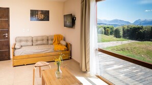 A room with a sofa, a wooden table, a vase with flowers, a painting on the wall, and a view of mountains through a window. - Mascardi Ground Floor Lake Front (Dina Huapi)