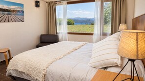 A bedroom with a bed, a chair, a lamp, and a view of a lake through the window.