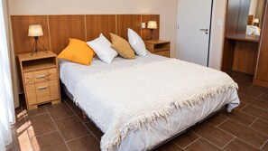 Habitación de hotel con una cama grande, cabecero de madera, dos mesitas de noche con lámparas, un armario y una puerta.