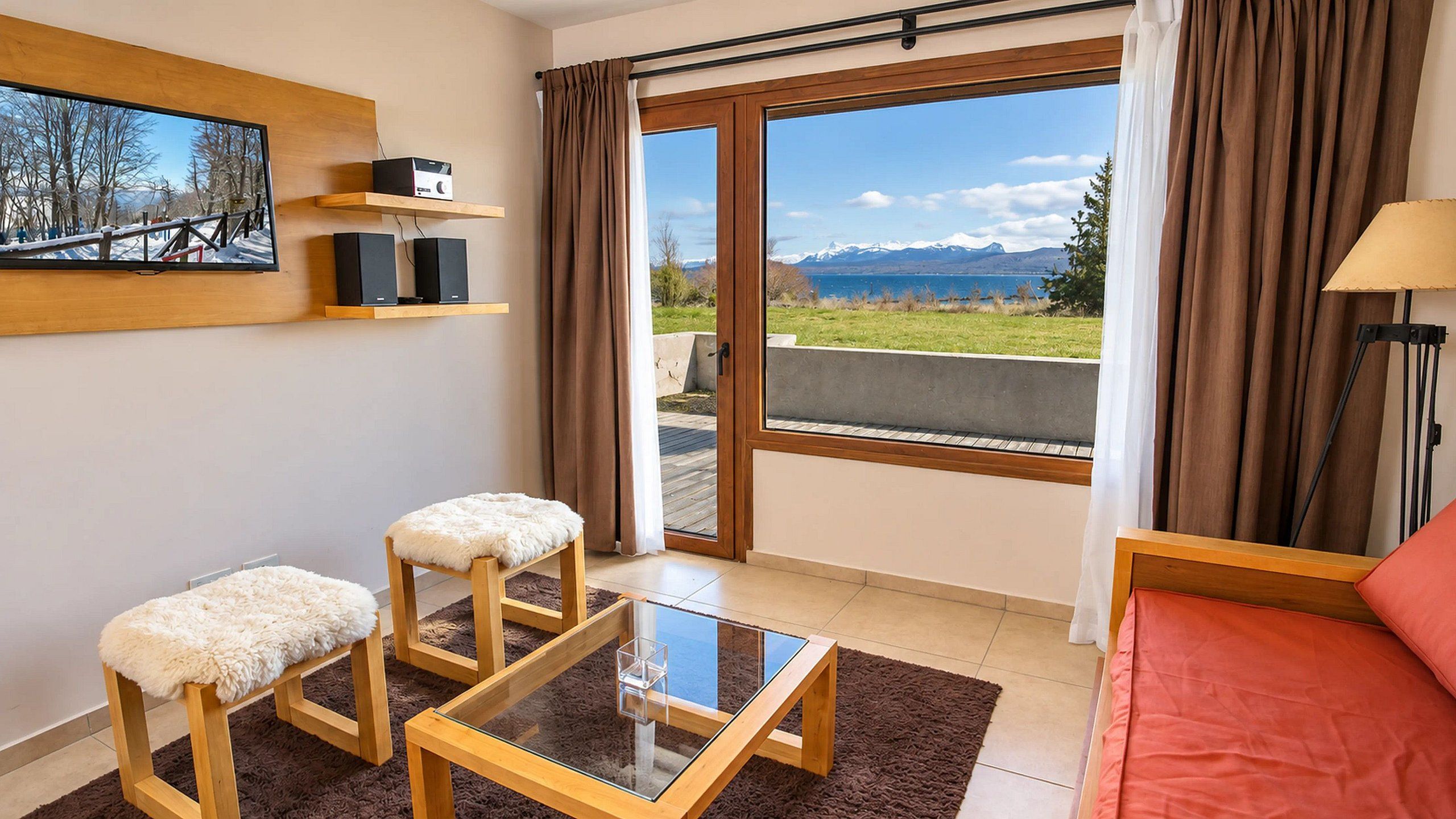 A modern room with a glass table, two cushioned stools, a sofa, and a view of mountains through a large window.