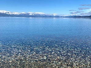 A clear lake with a pebbly shore and snow-capped mountains in the background.