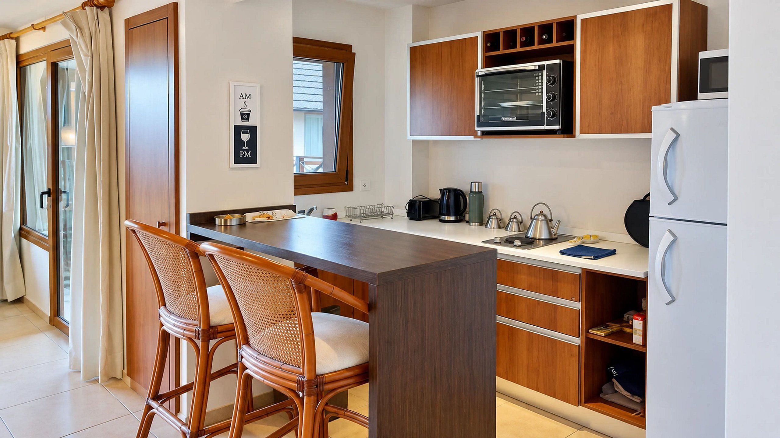 A compact kitchen with a central island, bar stools, a microwave, a kettle, and a refrigerator.