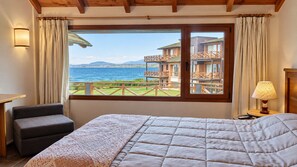 Una camera d'hotel con un letto grande, una sedia, un comodino con una lampada e una finestra con vista su un edificio e il mare.
