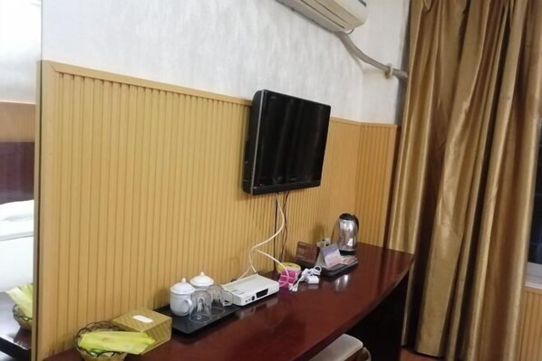 A hotel room with a wooden panel wall, a mounted television, a desk with a kettle, a tray with a box of tissues, and a window with curtains. - Wusheng Chengbei Business Hotel (Wusheng)