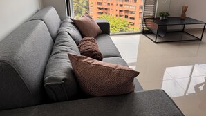 Een moderne woonkamer met een grijze bank, meerdere kussens, een kleine tafel en een uitzicht op een stadsbeeld door grote ramen.