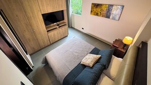 Een moderne slaapkamer met een groot bed, een tv bevestigd aan een houten kast, een schilderij aan de muur en een raam dat natuurlijk licht binnenlaat.