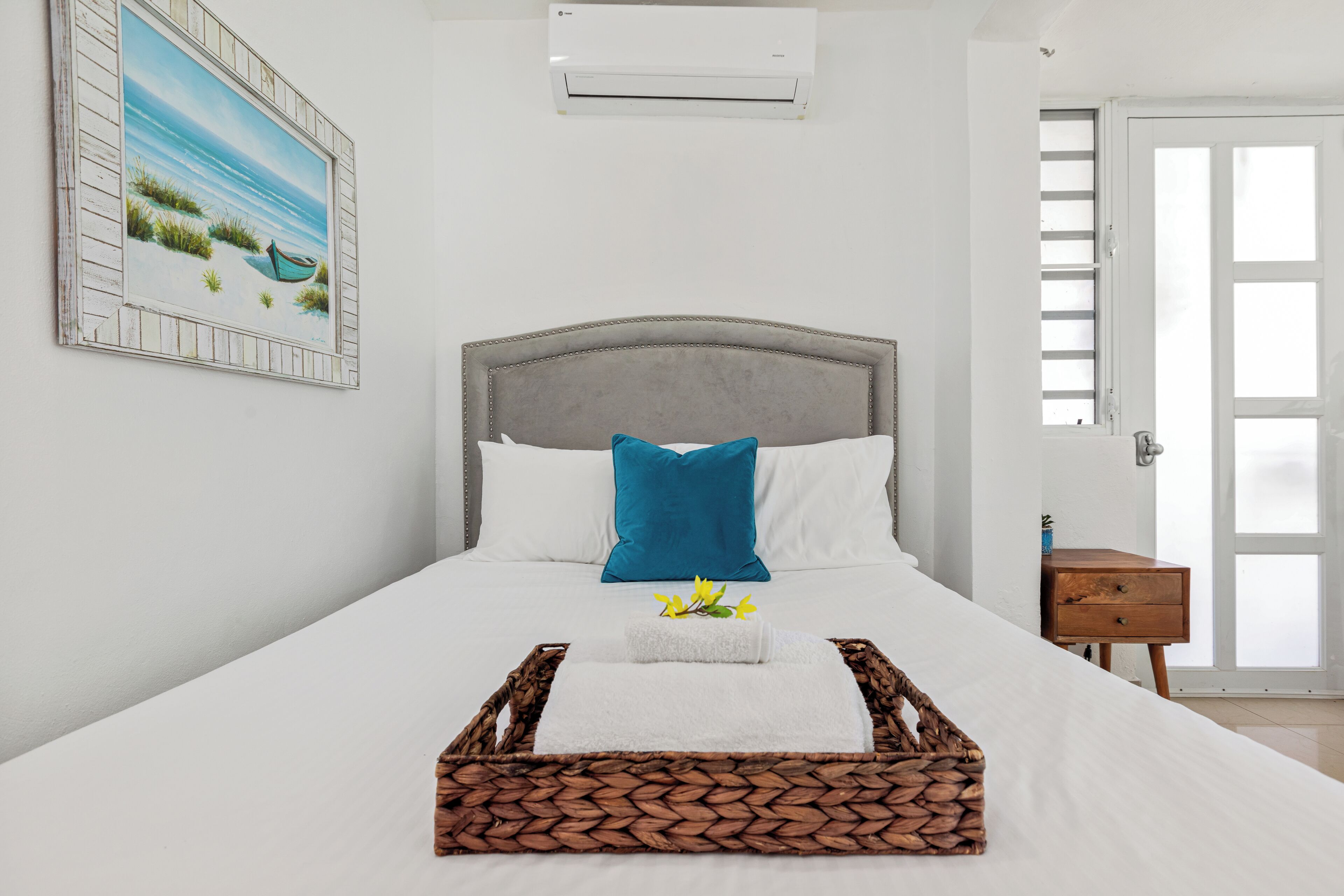 A neatly made bed with a headboard, a small wooden nightstand, a wicker tray, and a painting of a beach scene on the wall.