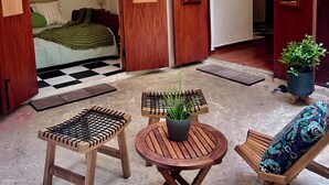 Une pièce avec un lit, une porte en bois, une petite table avec une plante et une chaise dotée d’un coussin à motifs.