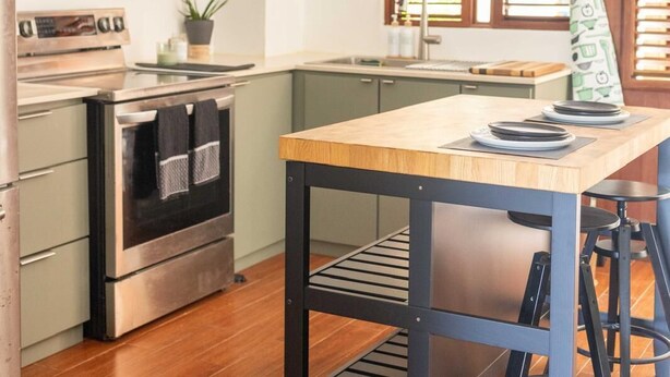 Une cuisine moderne avec une table en bois, un four encastré et un évier. On trouve des étagères au-dessus du plan de travail et une fenêtre avec des stores.
