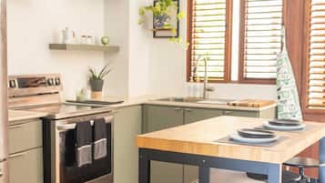 Een moderne keuken met een houten tafel, een inbouwoven en een gootsteen. Boven het aanrecht zijn planken en er is een raam met zonwering.