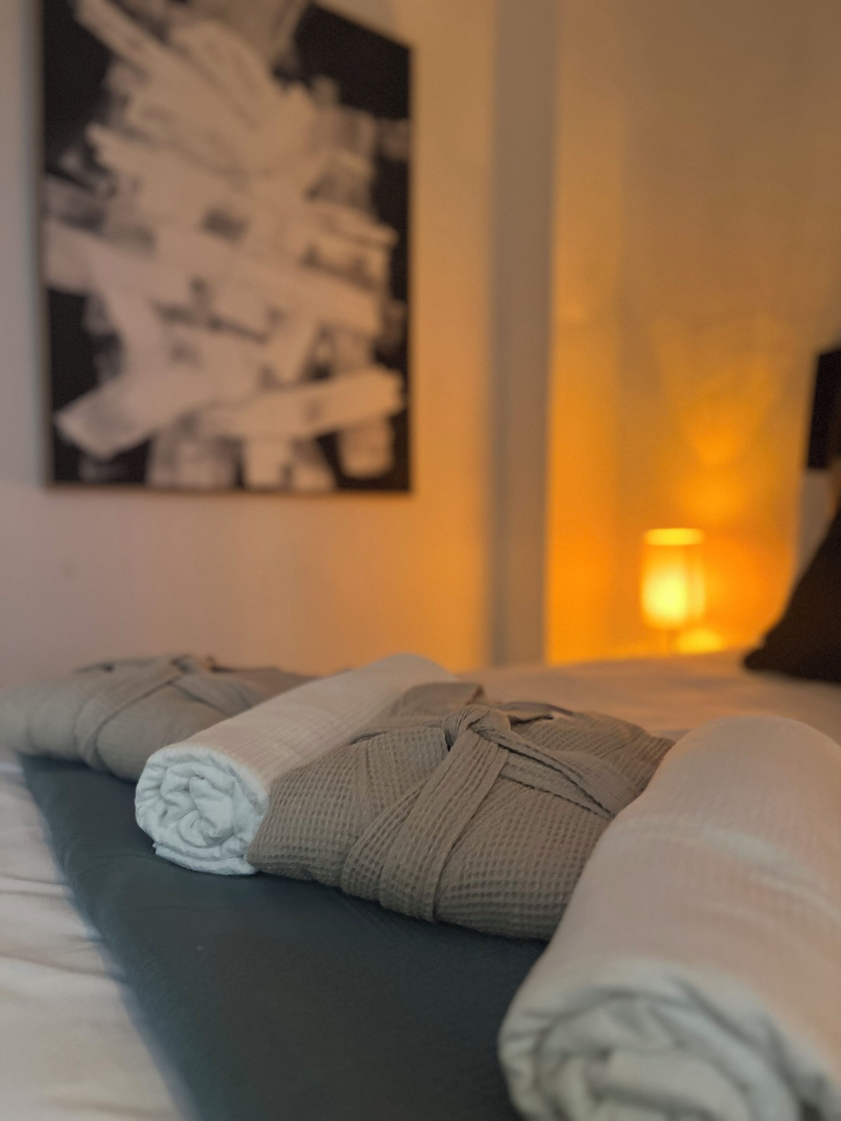A neatly made bed with folded towels, a wall-mounted artwork, and a lit bedside lamp.