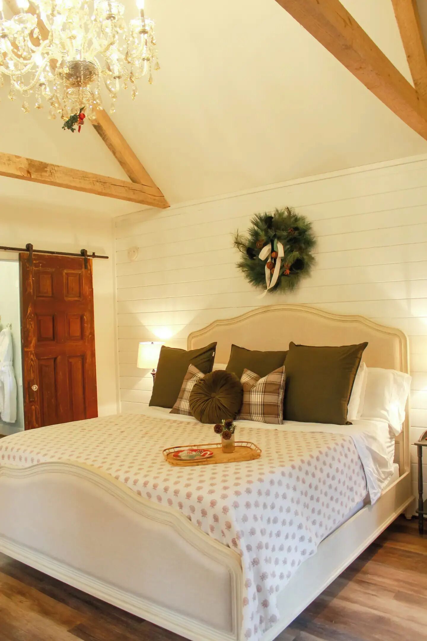A bedroom with a wooden ceiling, a chandelier, a bed with patterned bedding, and a Christmas wreath on the wall.