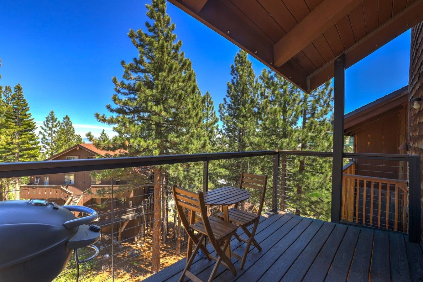 A wooden deck with a grill, a table, and chairs, overlooking a forested area.