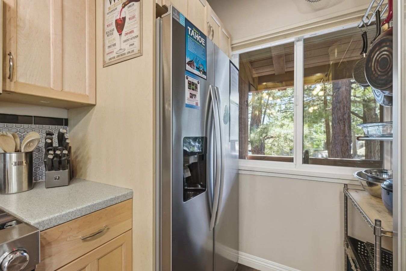 A kitchen with a stainless steel refrigerator, wooden cabinets, a window with a view of trees, and a metal rack with pots and pans.